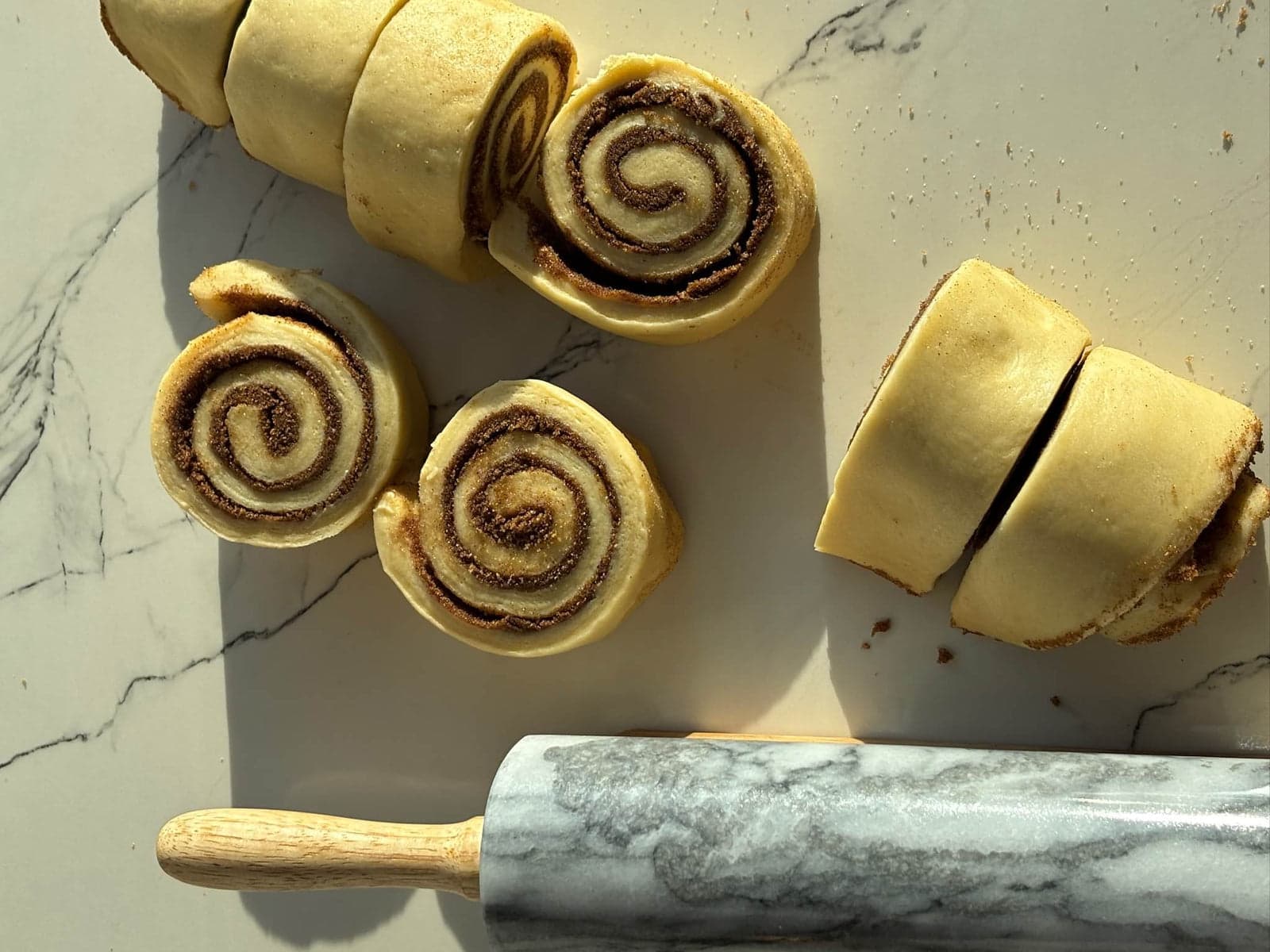 Sliced spirals of raw cinnamon roll dough beside a marble rolling pin